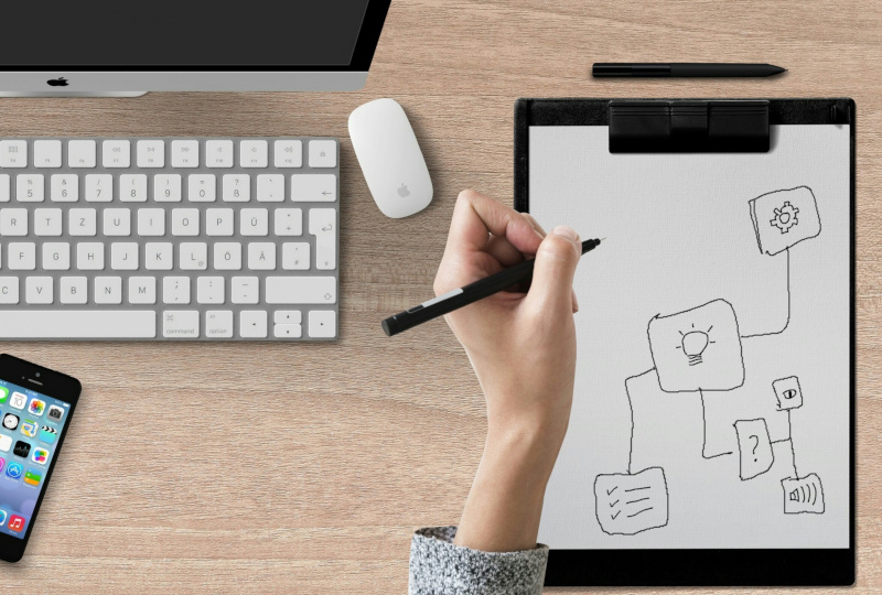 A left-handed person scribbling on a paper with clipboard while in front of a computer.